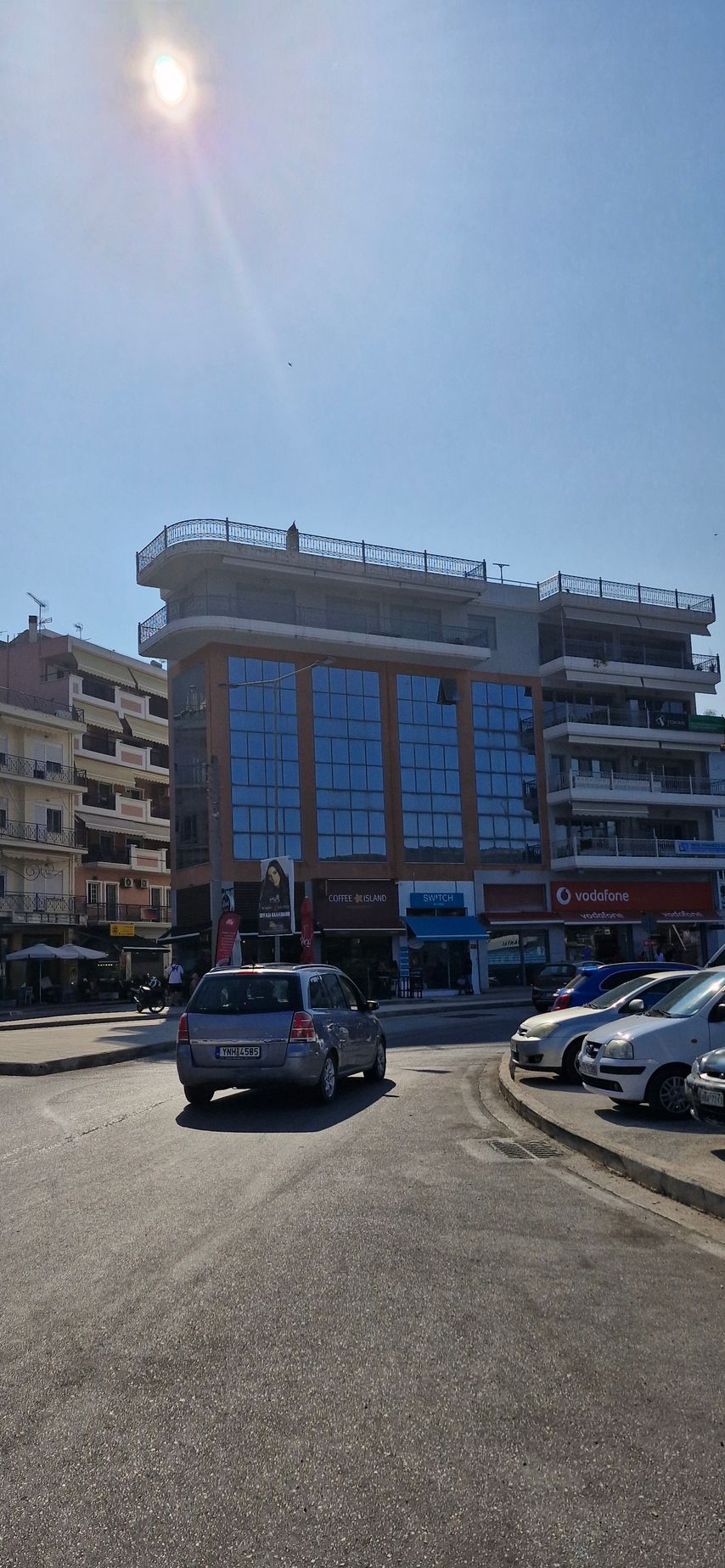 Ένα πολυώροφο κτίριο με γυάλινα παράθυρα, ένα αυτοκίνητο οδηγεί στο δρόμο με άλλα παρκαρισμένα αυτοκίνητα μια ηλιόλουστη μέρα.