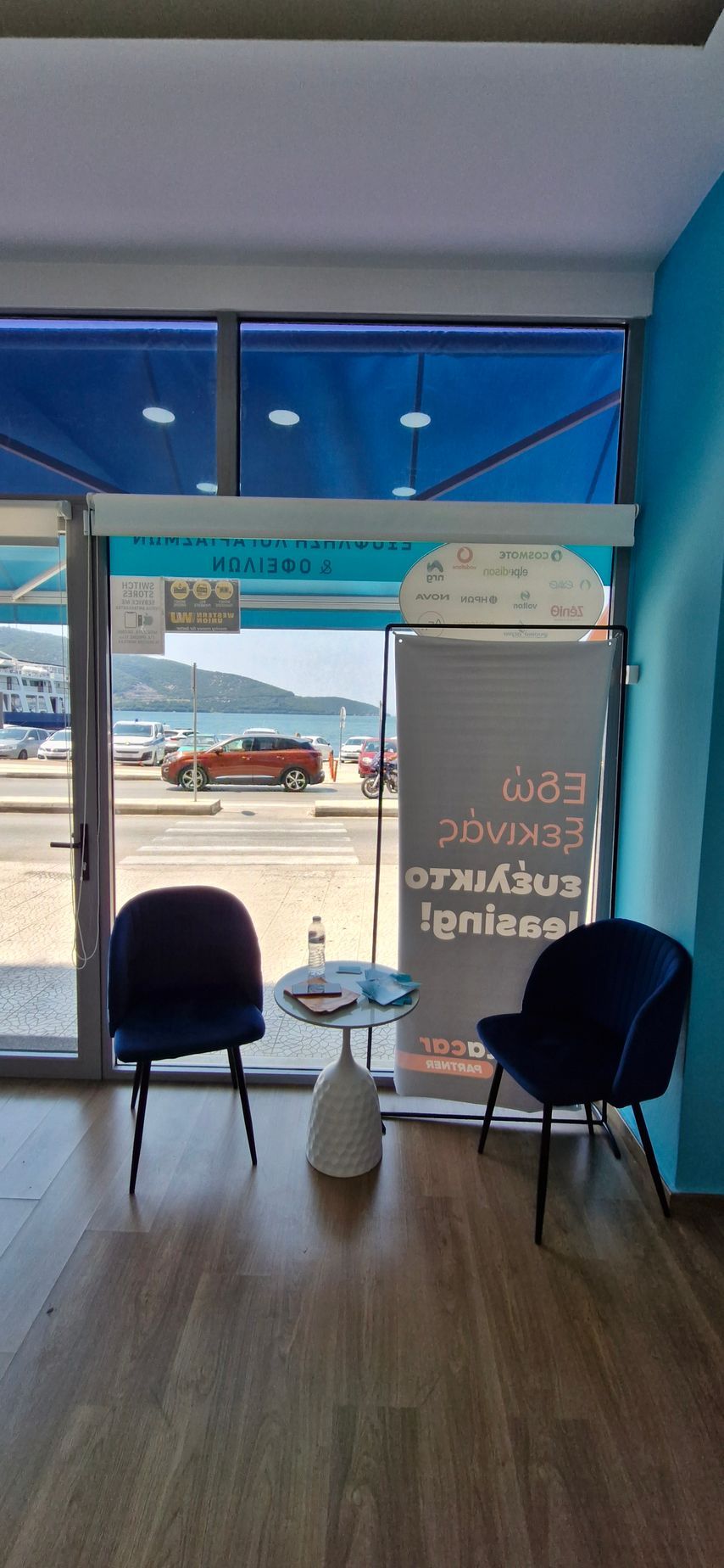 Ένα εσωτερικό με δύο καρέκλες σε μπλε σκούρο χρώμα και ένα μικρό τραπέζι που βλέπει σε ένα μεγάλο παράθυρο με θέα σε έναν δρόμο και τον ωκεανό.