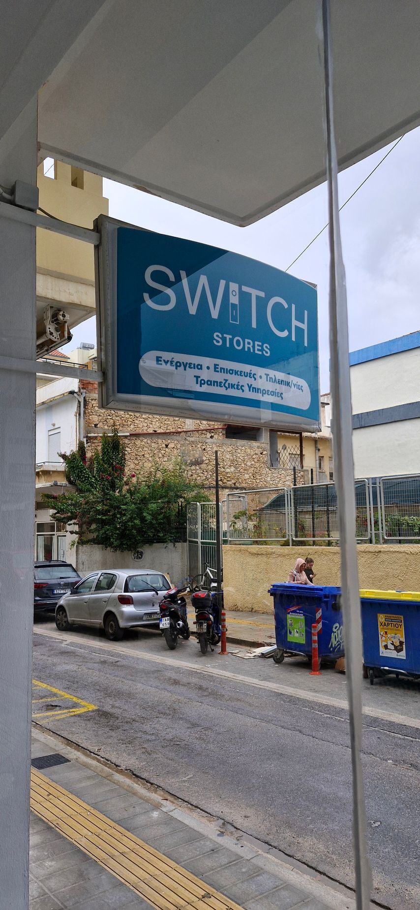 Πινακίδα για το Switch Sports, μπλε με λευκό κείμενο, σε έναν δρόμο με αυτοκίνητα, κάδους απορριμμάτων και ένα μερικώς ορατό κτίριο.