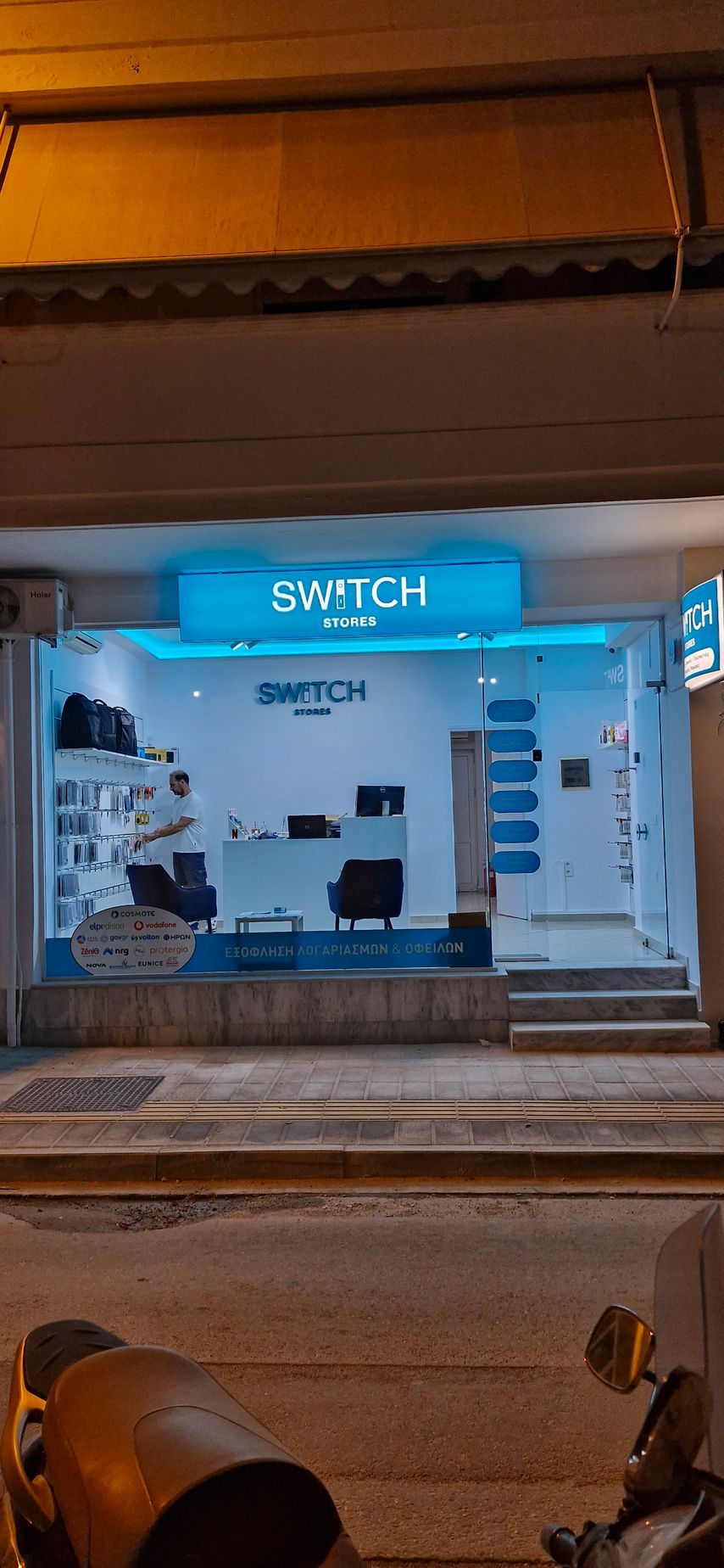 Εξωτερικό ενός καταστήματος Switch τη νύχτα, με φωτεινή μπλε πινακίδα, βιτρίνα καταστήματος και μια μοτοσικλέτα.