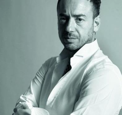 Άνδρας με λευκό πουκάμισο με σταυρωμένα χέρια, κοιτάζει την κάμερα. Ασπρόμαυρη φωτογραφία.