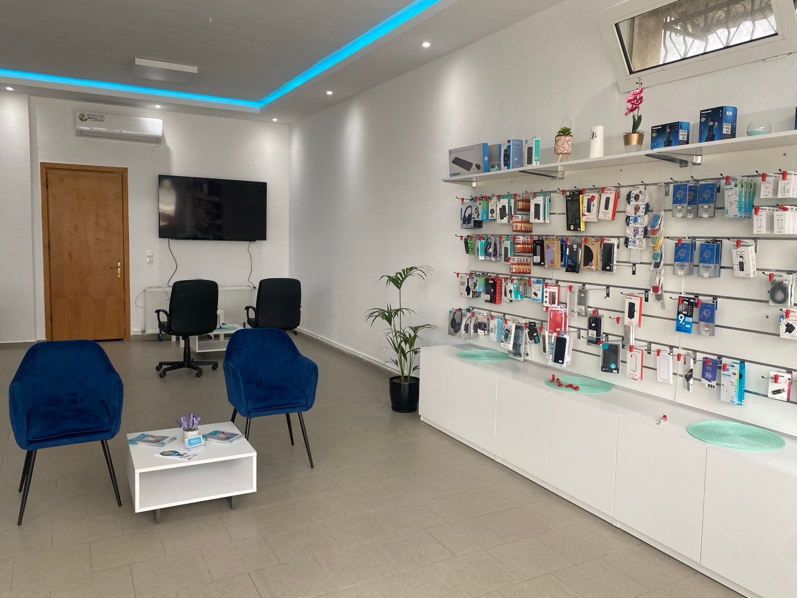 Εσωτερικό ενός καταστήματος κινητής τηλεφωνίας με εκθέσεις προϊόντων σε λευκά ράφια, χώρος αναμονής με μπλε καρέκλες.