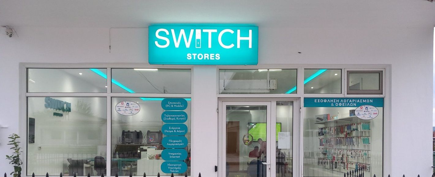 Εξωτερικό των καταστημάτων Switch με τιρκουάζ πινακίδες. Διαφανή γυάλινα παράθυρα δείχνουν το εσωτερικό ενός καταστήματος με εμπορεύματα.
