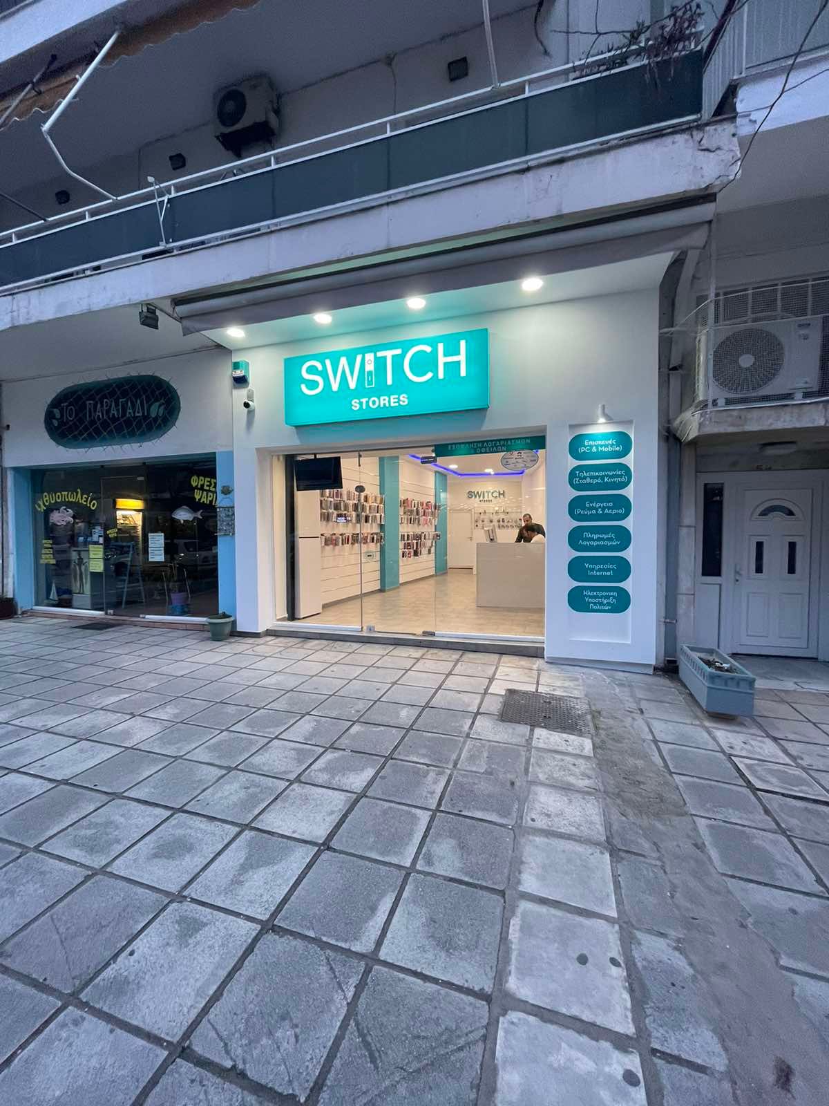 Εξωτερικό ενός καταστήματος Switch με το σήμα του τιρκουάζ, με ένα μικρό κατάστημα, λευκούς τοίχους και πλακόστρωτο πεζοδρόμιο.