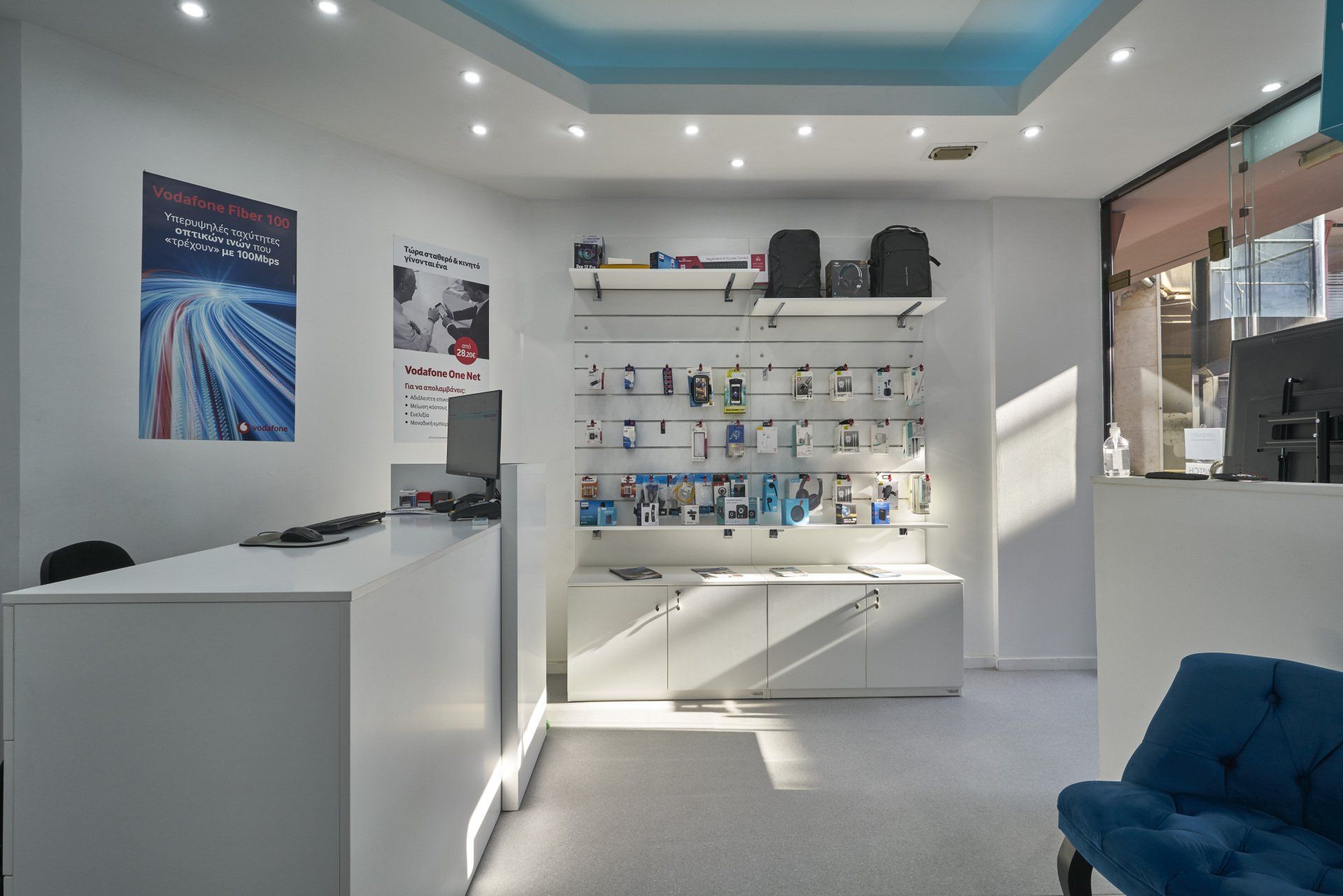 Εσωτερικό ενός συνεργείου επισκευής τηλεφώνων με λευκούς τοίχους, ράφια προβολής, έναν πάγκο και μια μπλε καρέκλα.