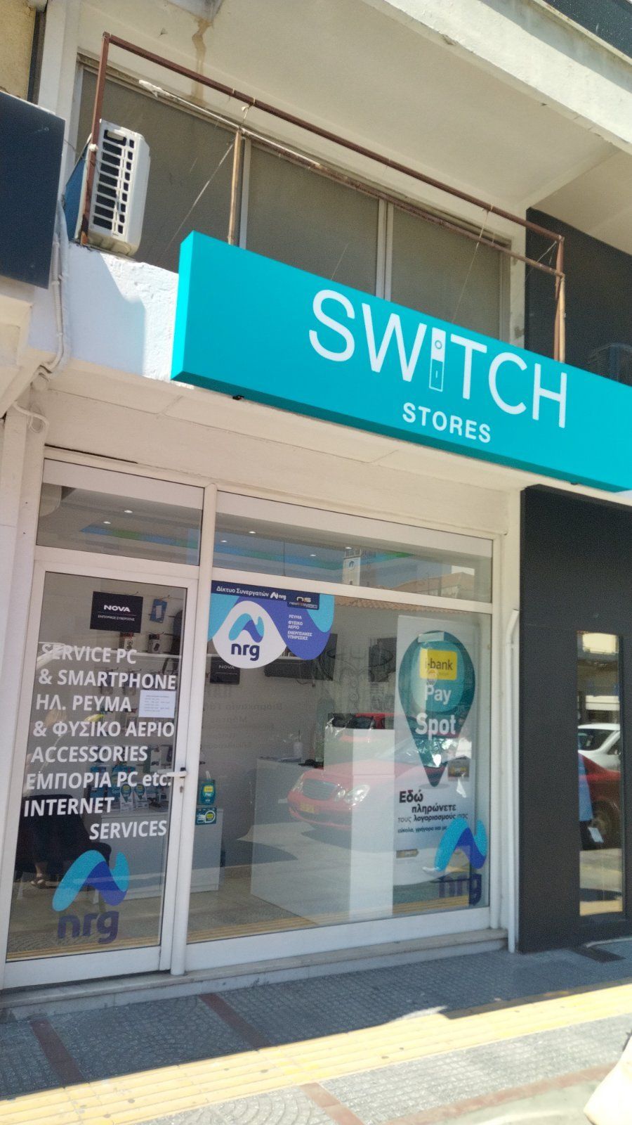 Εξωτερικό καταστήματος Switch με γαλαζοπράσινη πινακίδα. Λευκή και γυάλινη βιτρίνα, με είδη τεχνολογίας και υπηρεσίες που προσφέρονται.