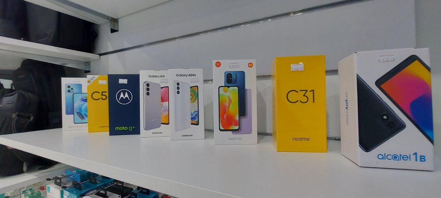 Μια σειρά από κουτιά smartphone που εκτίθενται σε ένα λευκό ράφι. Τα κουτιά είναι διαφόρων χρωμάτων, εμπορικών σημάτων και μεγεθών.