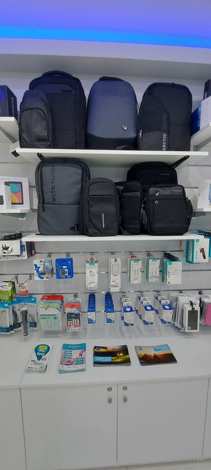 Μια βιτρίνα καταστήματος με σακίδια πλάτης στα ράφια, διάφορα αξεσουάρ από κάτω. Λευκά ντουλάπια και τοίχοι, μπλε φως.