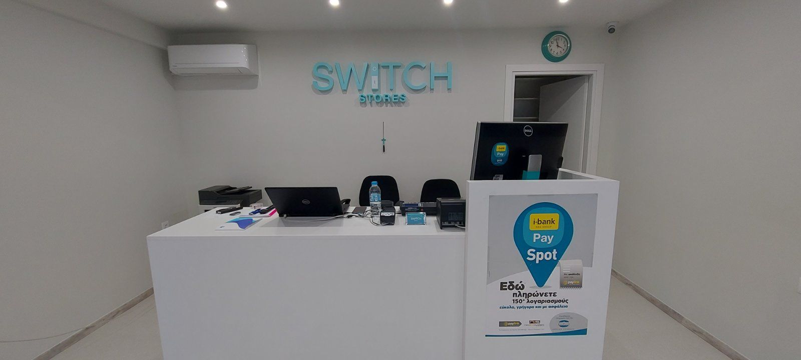 Εσωτερικό ενός καταστήματος Switch με λευκό πάγκο, γαλαζοπράσινη πινακίδα και ανοιχτή πόρτα.