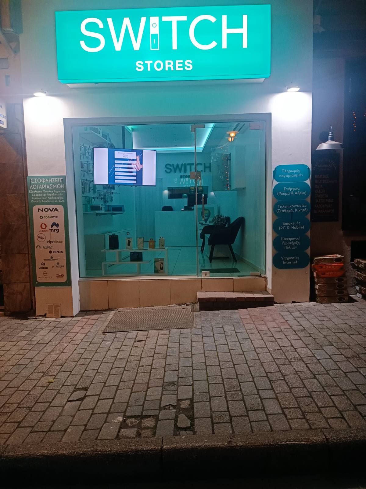 Η βιτρίνα του καταστήματος Switch Stores φωτιζόταν τη νύχτα, με προϊόντα σε έκθεση, γαλαζοπράσινες πινακίδες και πεζοδρόμιο στρωμένο με τούβλα.