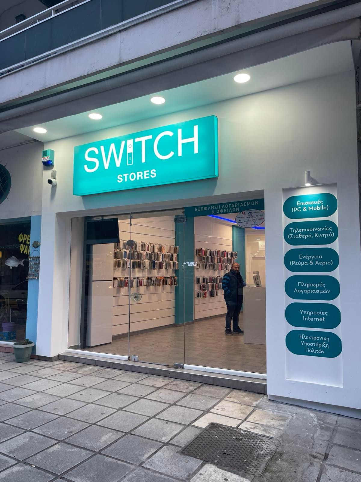«Εξωτερική όψη των καταστημάτων Switch. Ένα άτομο στέκεται στην πόρτα, κάτω από μια γαλαζοπράσινη πινακίδα. Λευκό κτίριο, γυάλινες πόρτες.»