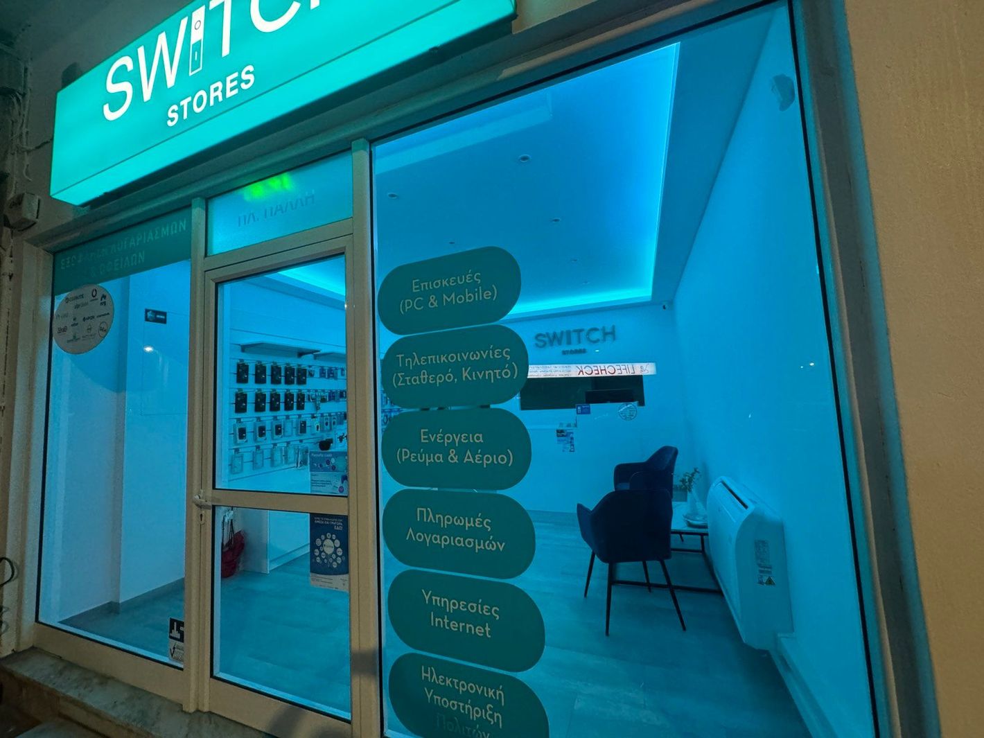 Βιτρίνα καταστήματος Switch Stores με μπλε τζάμια και φωτεινή επιγραφή. Υπηρεσίες που αναγράφονται στο τζάμι.