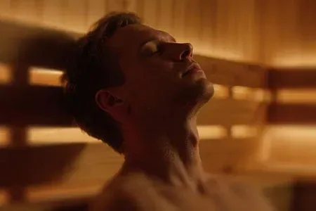 A person with eyes closed leans their head back, relaxing against the wood-paneled walls of a dimly lit sauna.