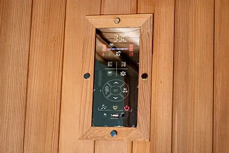 A digital sauna control panel mounted on wood-paneled walls with temperature and setting icons displayed on the screen.