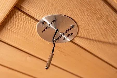 A metal temperature sensor mounted to wooden sauna ceiling panels.