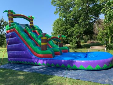A dual-lane, purple and green inflatable tropical water slide with palm tree decorations on a grassy lawn.