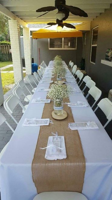A long table set for an event under a covered patio, featuring a burlap runner, baby's breath centerpieces, and white chairs.