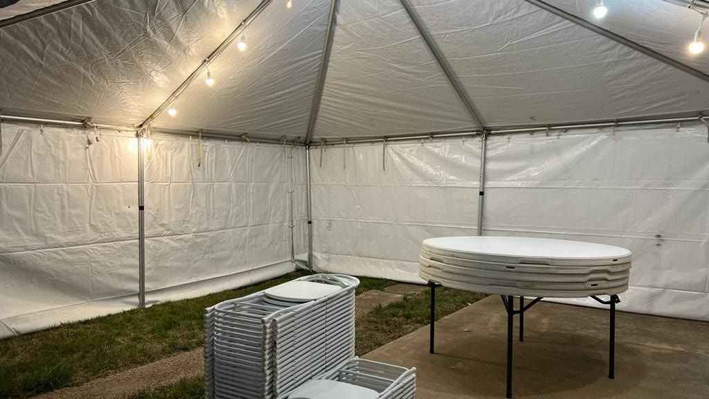 Inside a white event tent at night, a circular folding table stands next to stacks of folded chairs on grass and concrete.