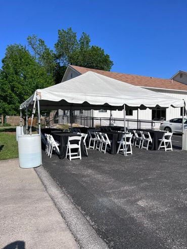 A white event tent set up on an asphalt lot with rows of black-clothed tables and white folding chairs.