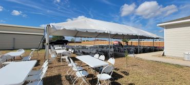 Outdoor event setup with white folding tables and chairs under a large white tent on a sunny day.
