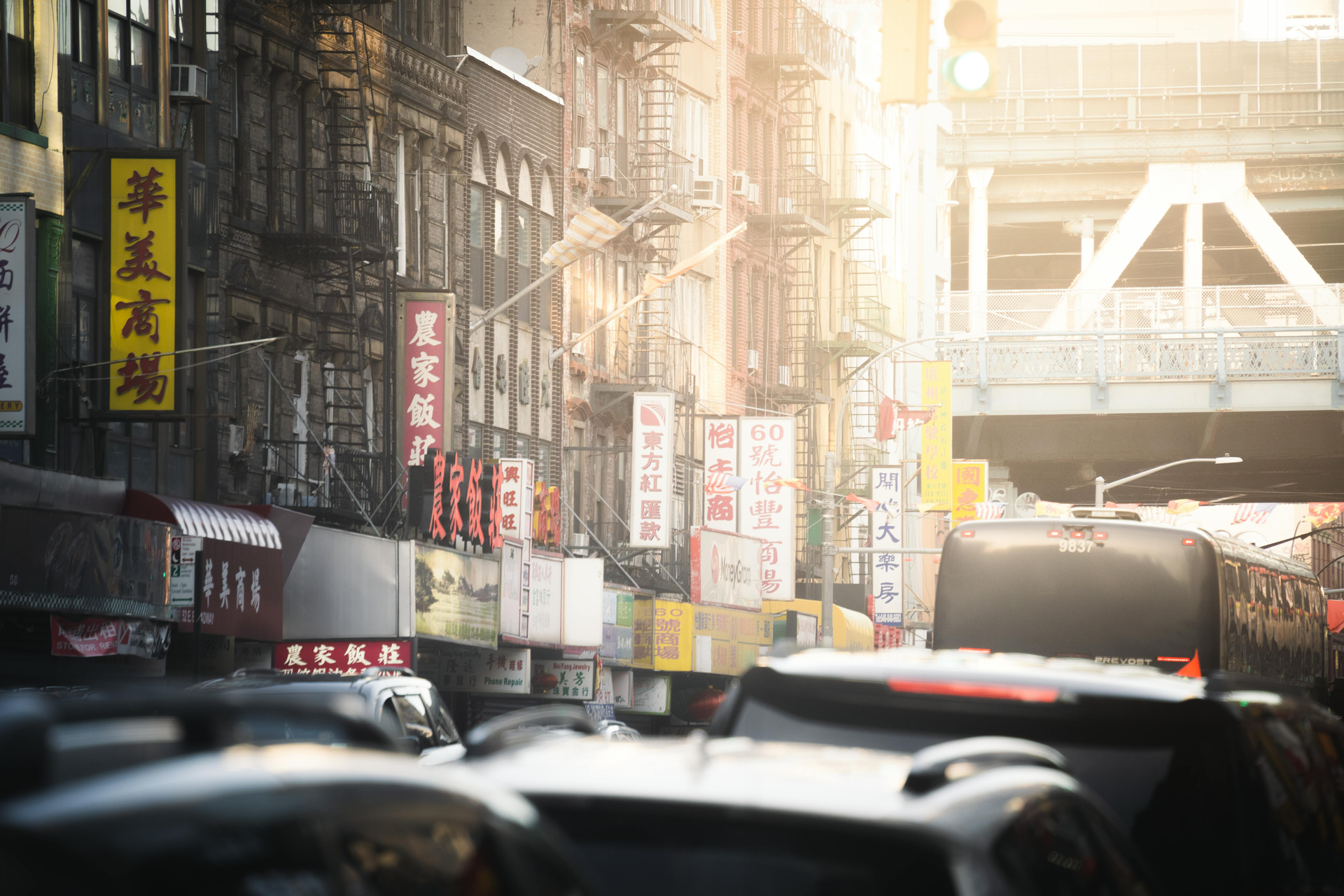 Busy urban street with traffic, storefront signs, and a sunlit pedestrian bridge in the distance