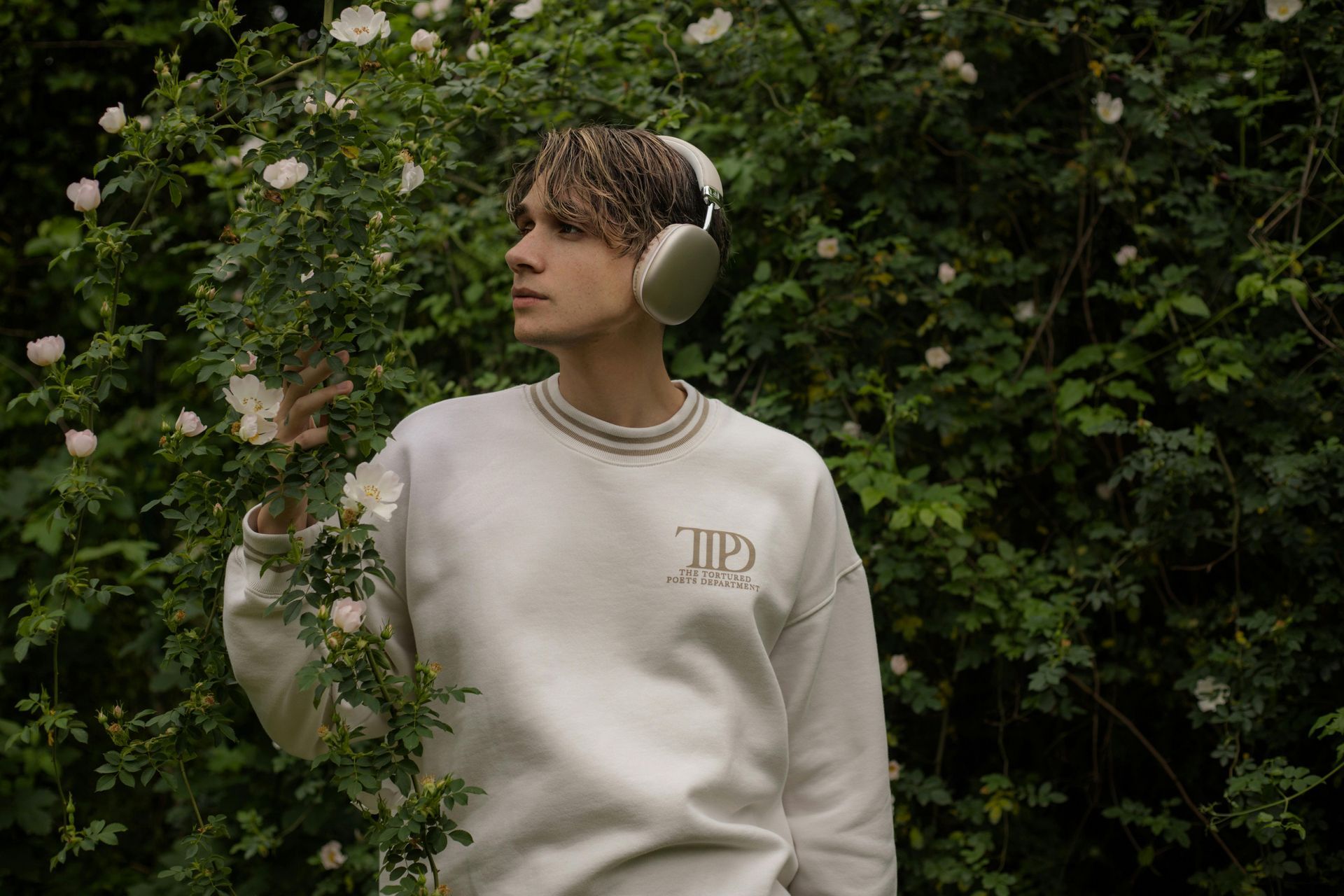 Man wearing headphones, white sweater, stands near a flowering bush, looking to the side.