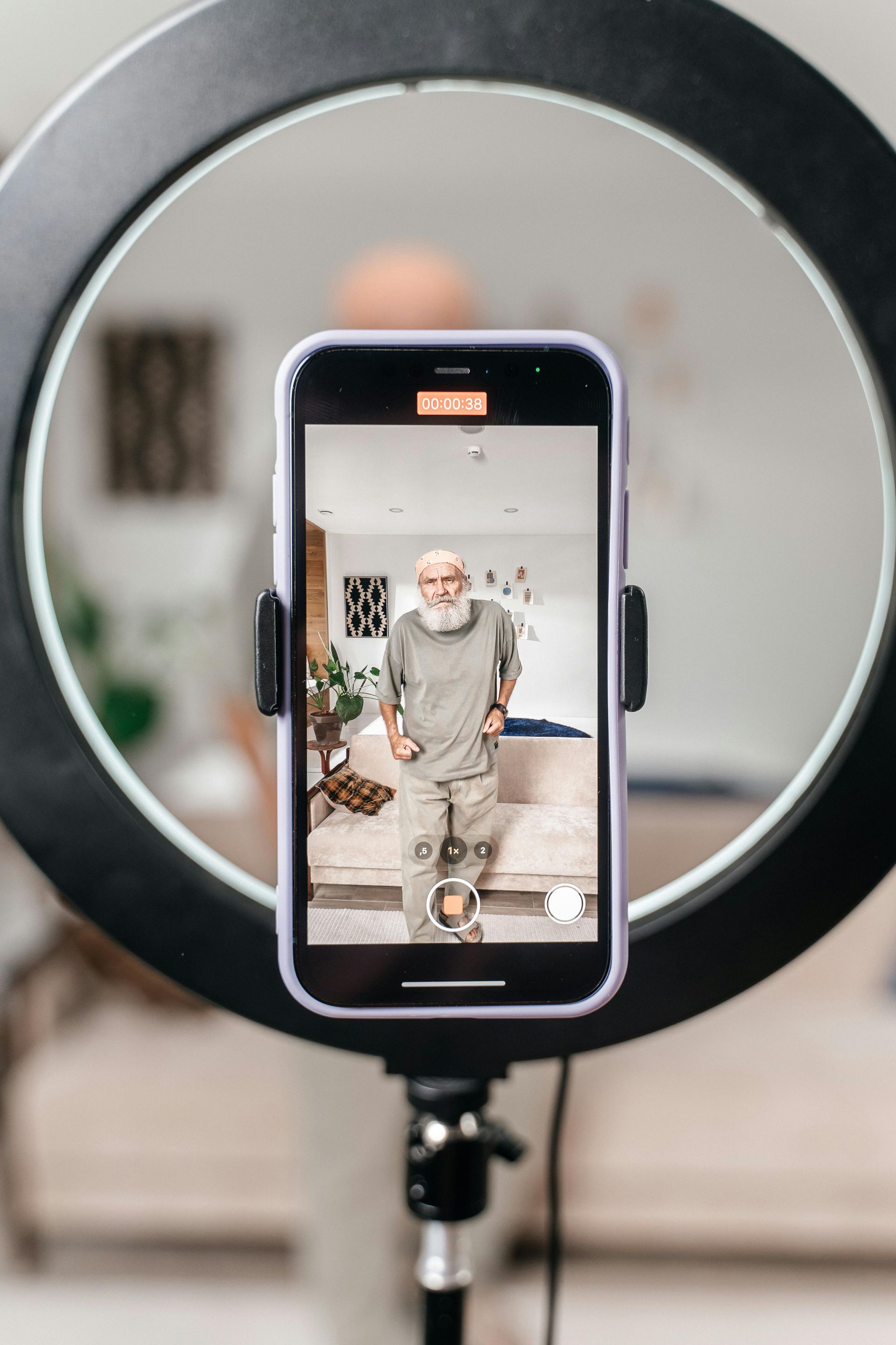 Phone on a ring light tripod recording a seated man in a living room studio setup.