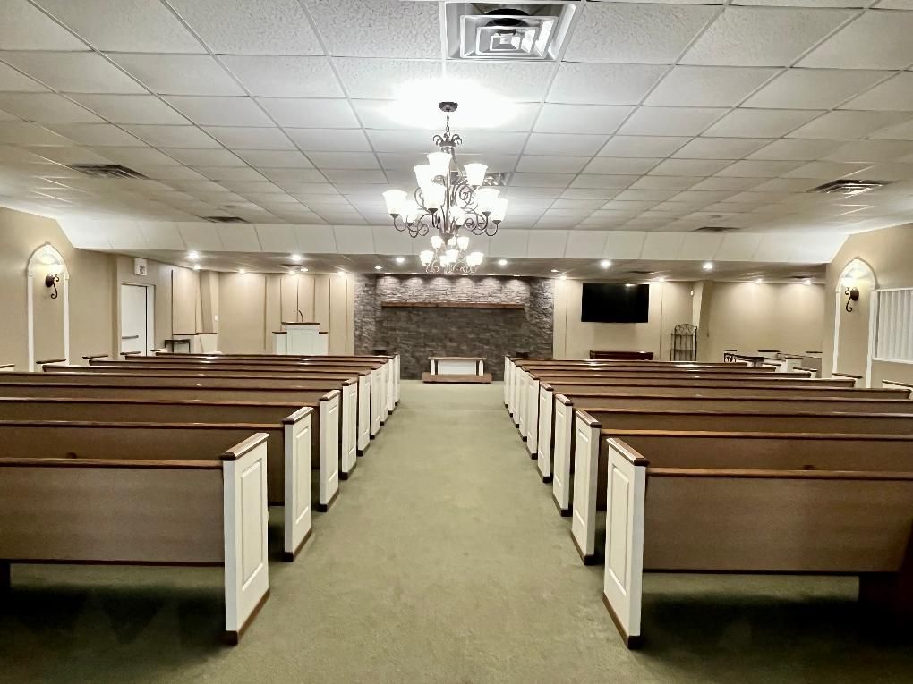 A chapel interior with rows of pews facing a raised platform, a chandelier, and a television.