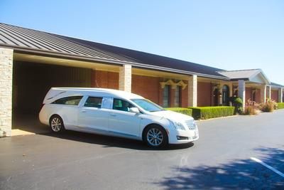 White hearse parked outside a brick building with a dark roof on a sunny day.