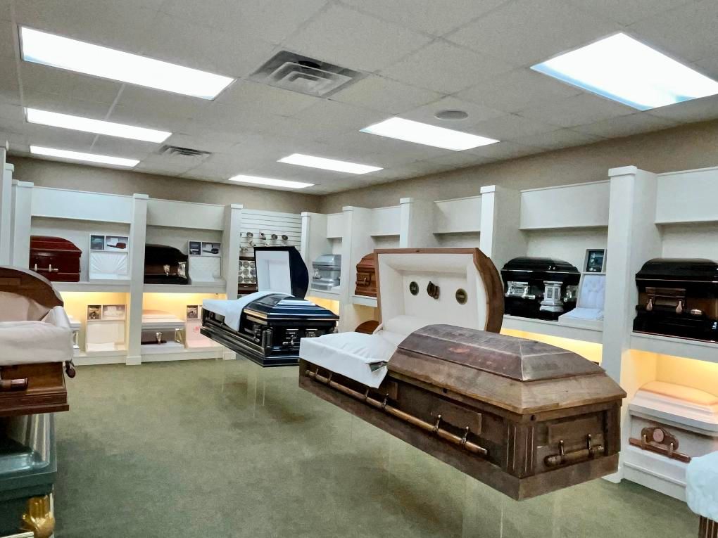 Coffin showroom with various caskets on display, lit by fluorescent lights.
