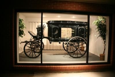 Black horse-drawn hearse displayed in a lit storefront window, flanked by potted plants.