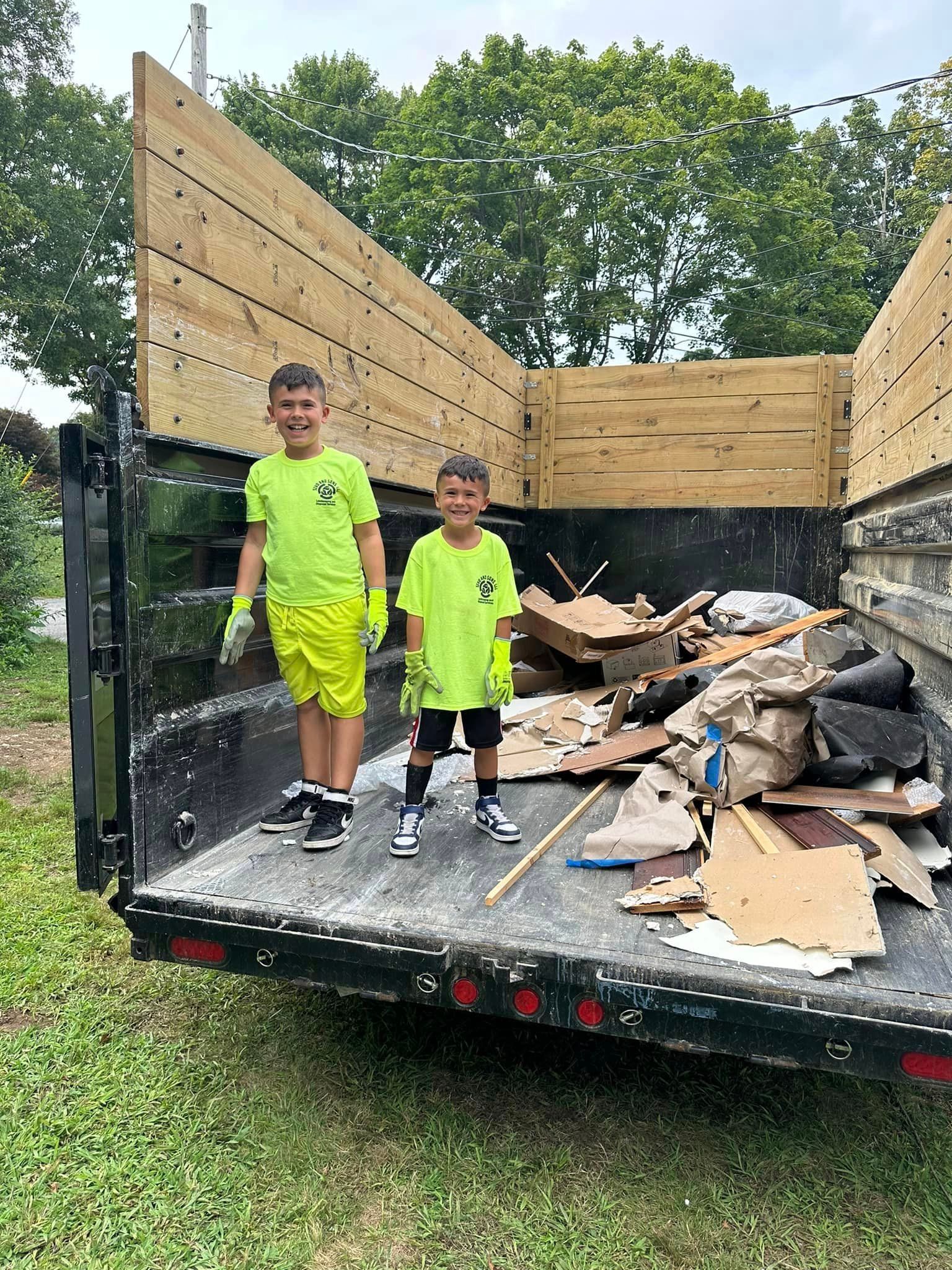 Two young boys are standing in the back of a dumpster filled with cardboard.