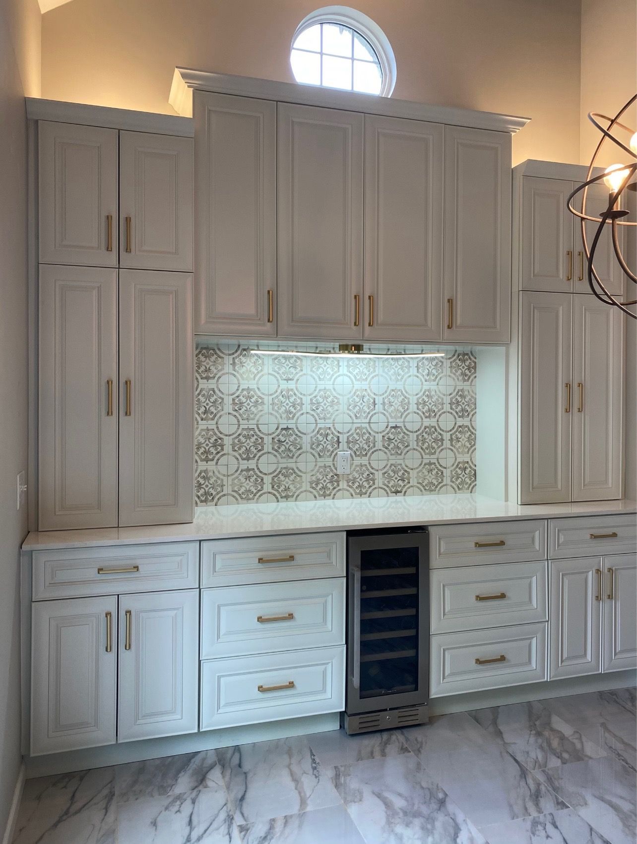 White built-in kitchen cabinets with patterned tile backsplash and marble-look floor under a small arched window.