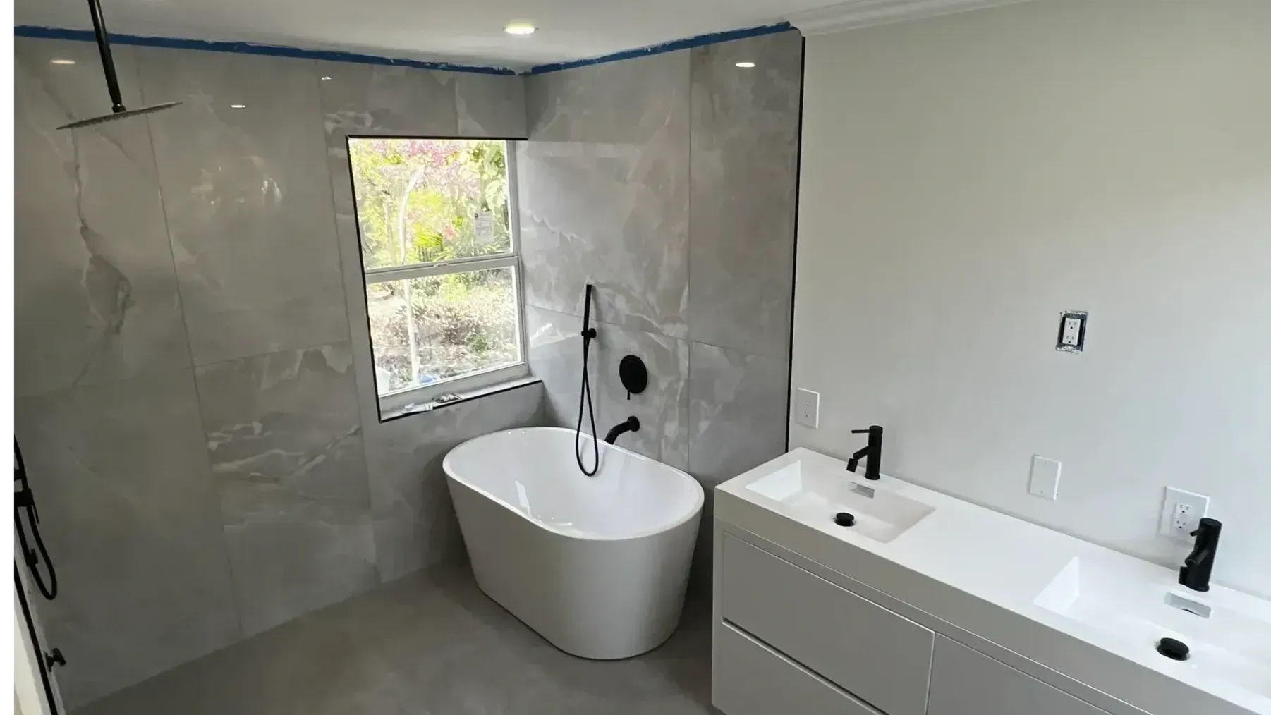 Modern bathroom with freestanding tub, double sink vanity, gray tile walls, and a window.