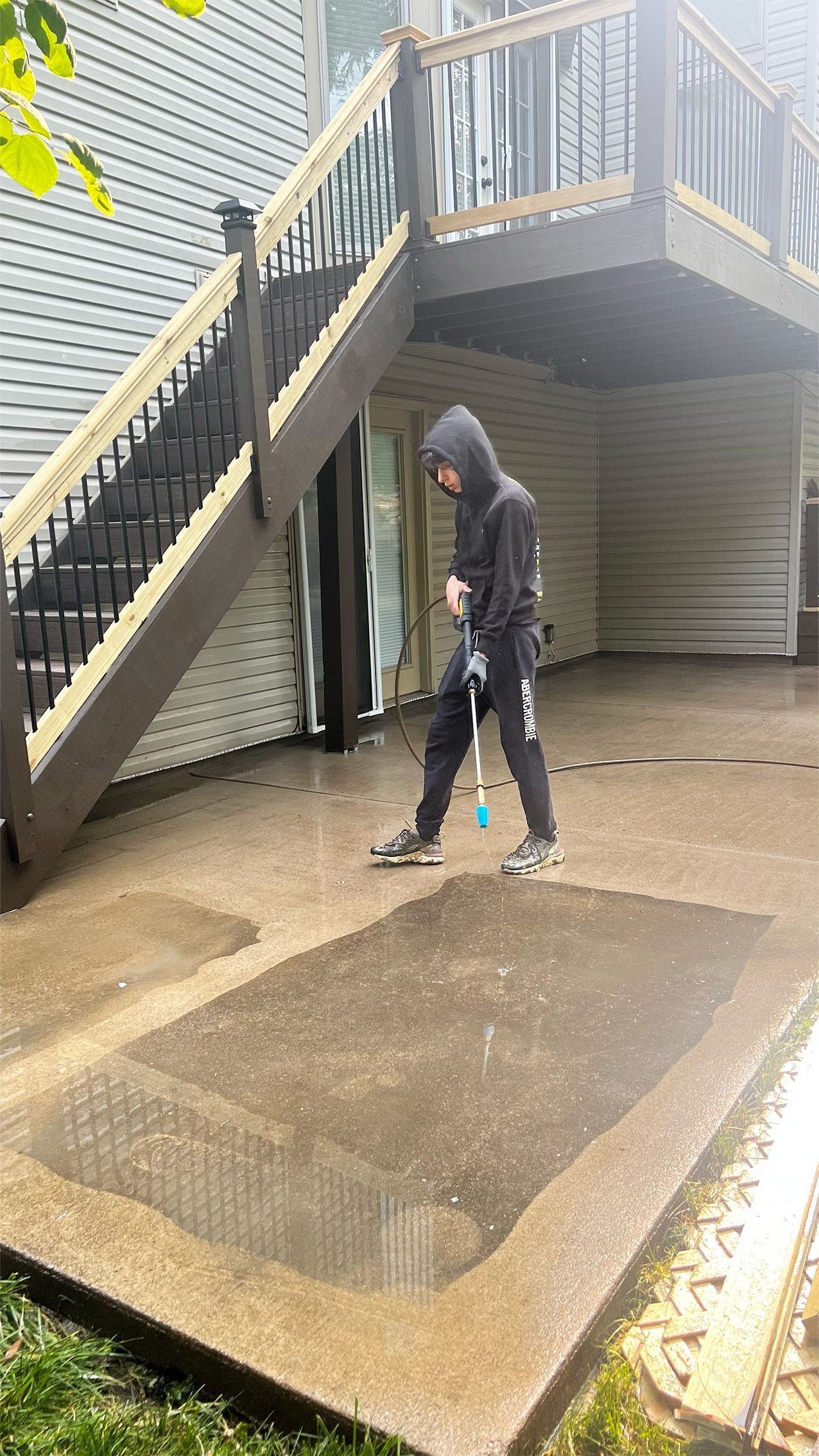 Person in a hooded jacket pressure-washing a wet concrete patio under outdoor stairs.