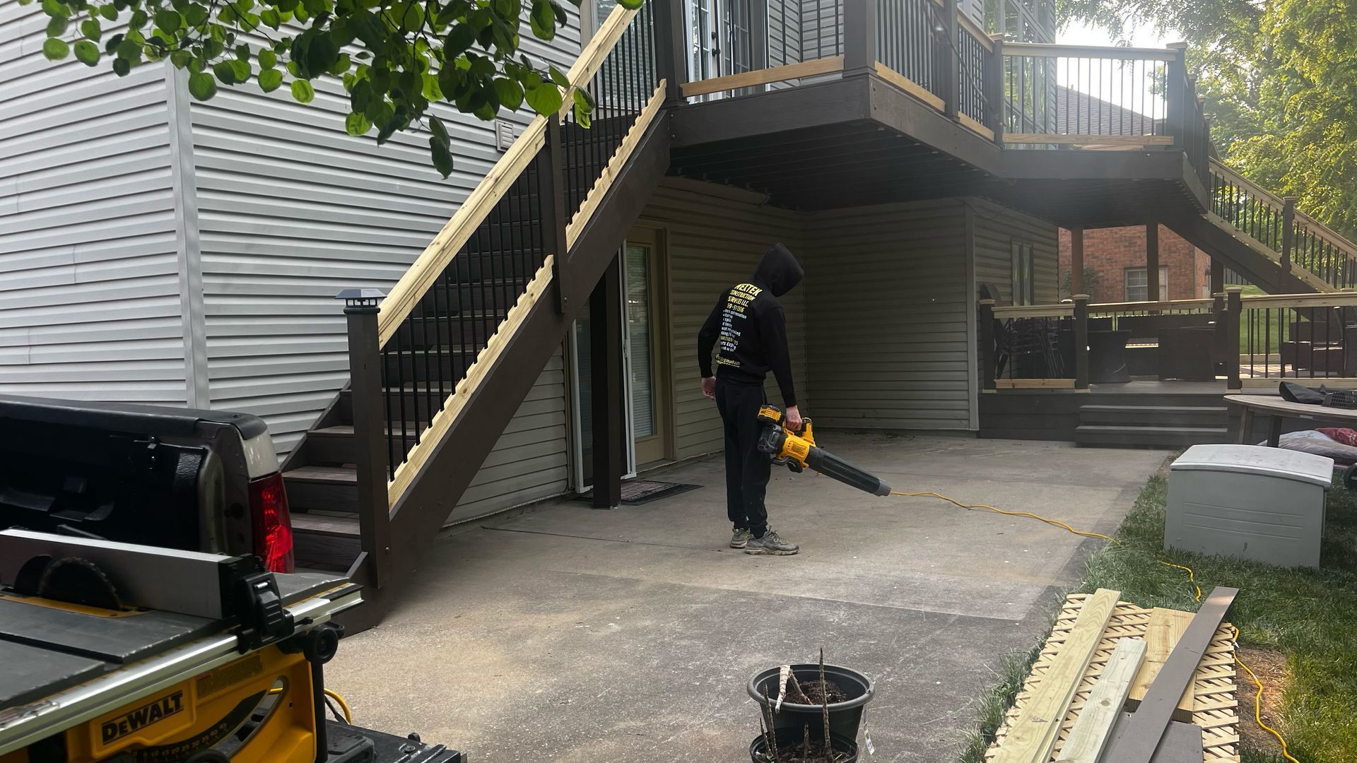 Worker using a leaf blower in a driveway beneath an outdoor staircase.