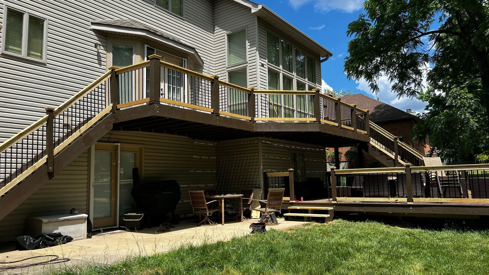 Wooden deck with stairs on a beige house, set in a sunny backyard with grass and trees.