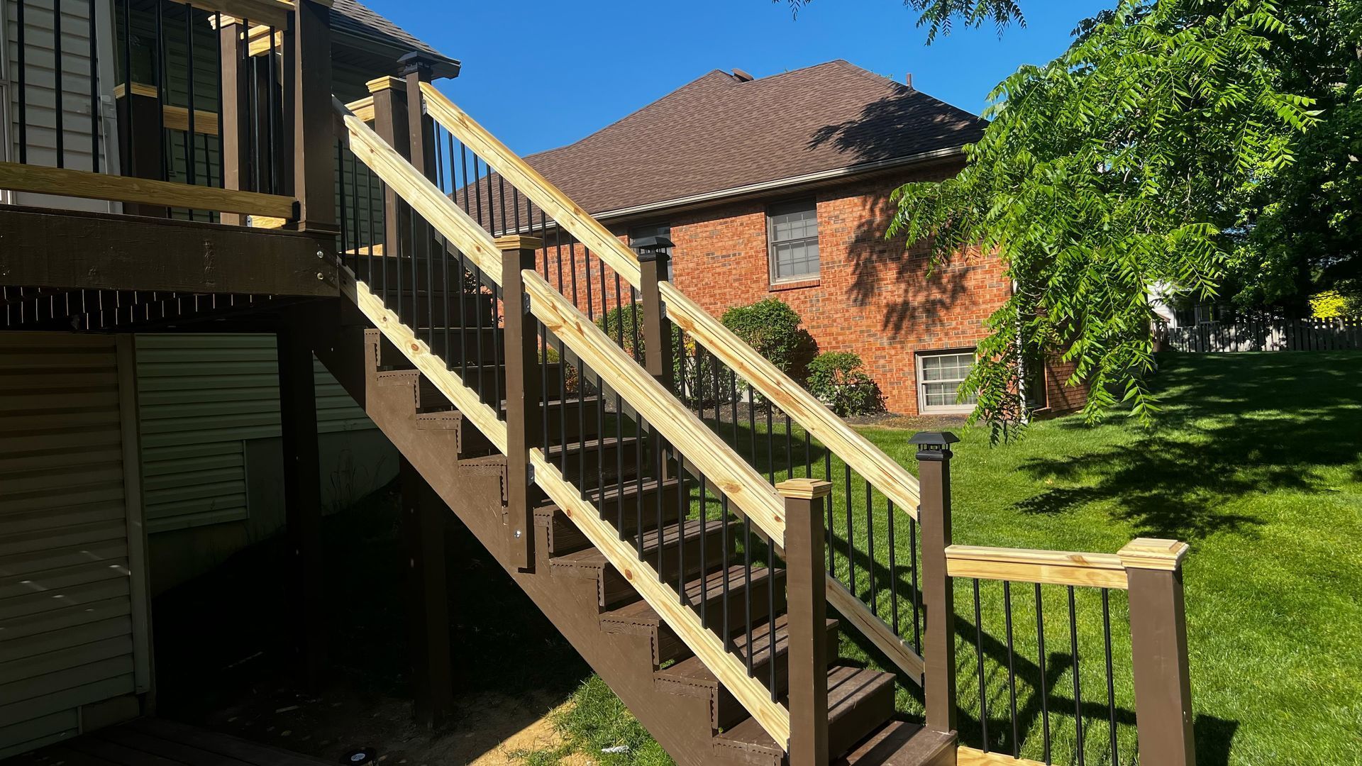 Wooden outdoor staircase beside a brick building, leading up from a shaded yard with green lawn.