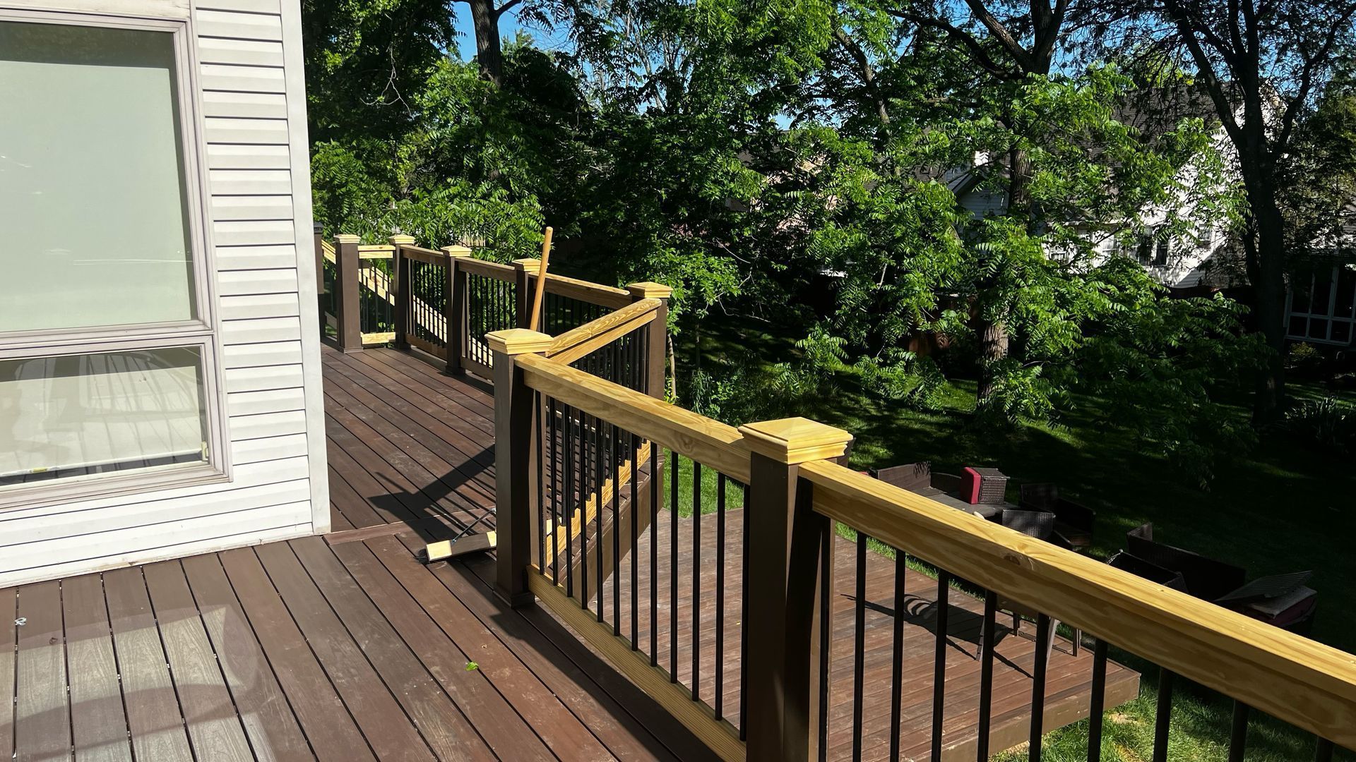 Wooden deck with railing beside a house, overlooking leafy trees and a shaded yard