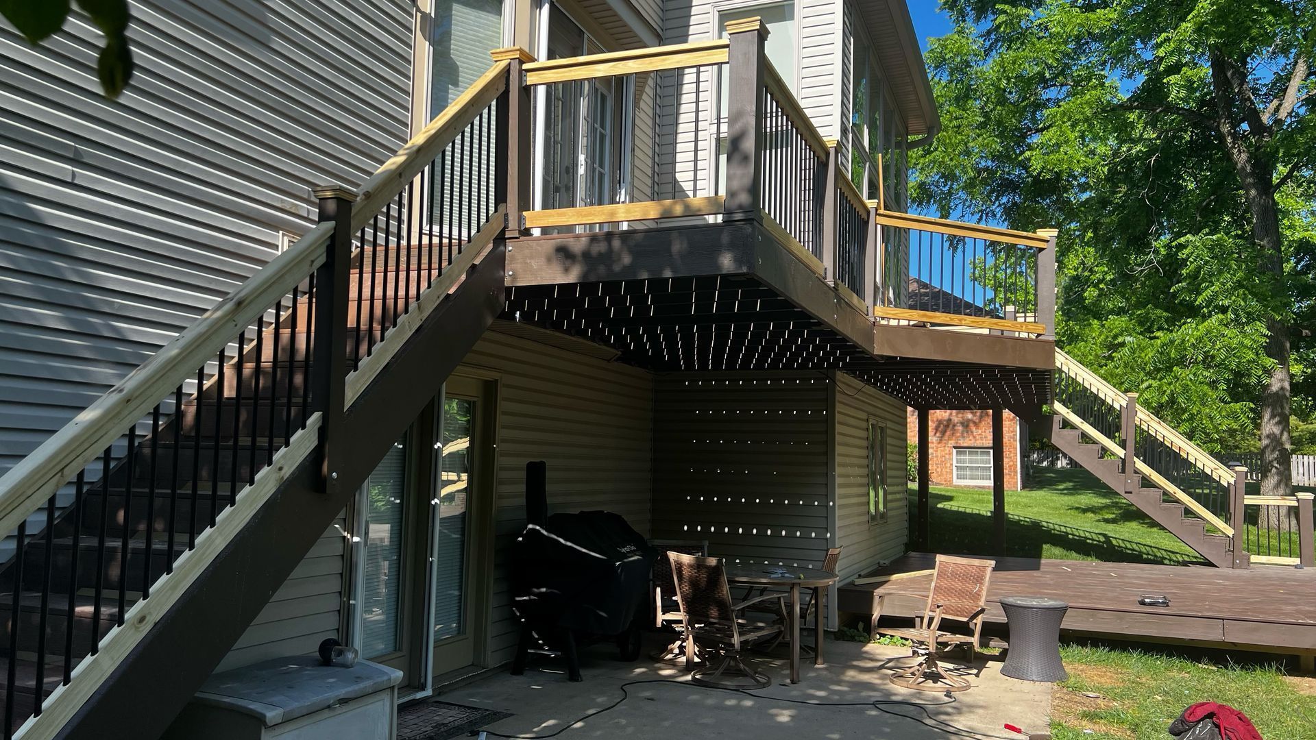 Backyard deck above a basement patio, with stairs, railing, and trees in bright sunlight