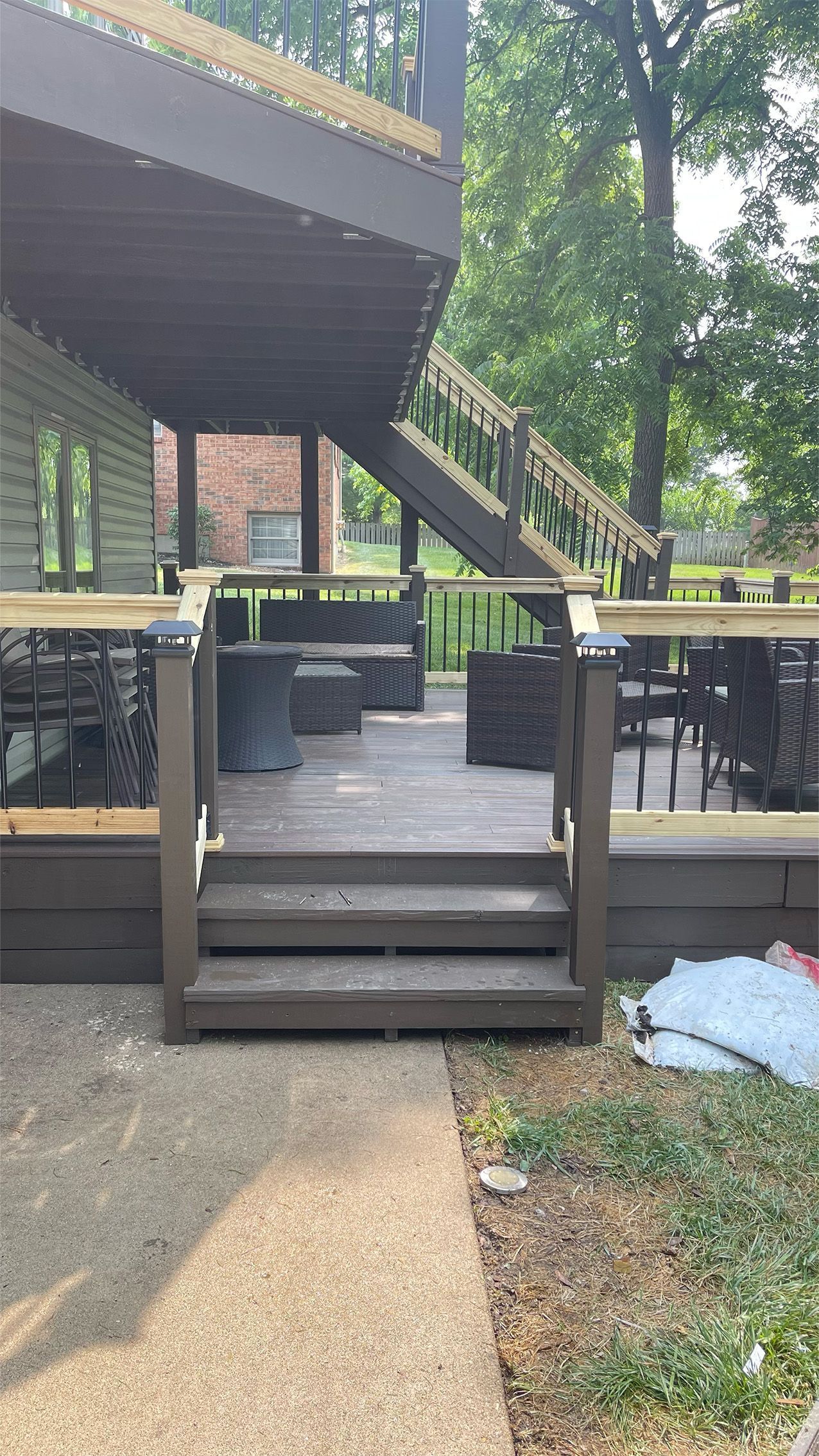 Outdoor deck steps with black railings leading to a raised patio beside a wooded house.