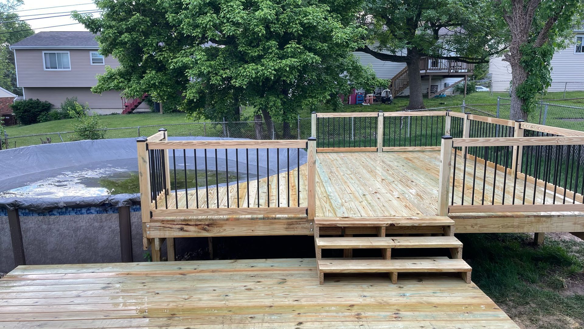 Wooden backyard deck under construction with railings and steps, backed by a covered pool and trees