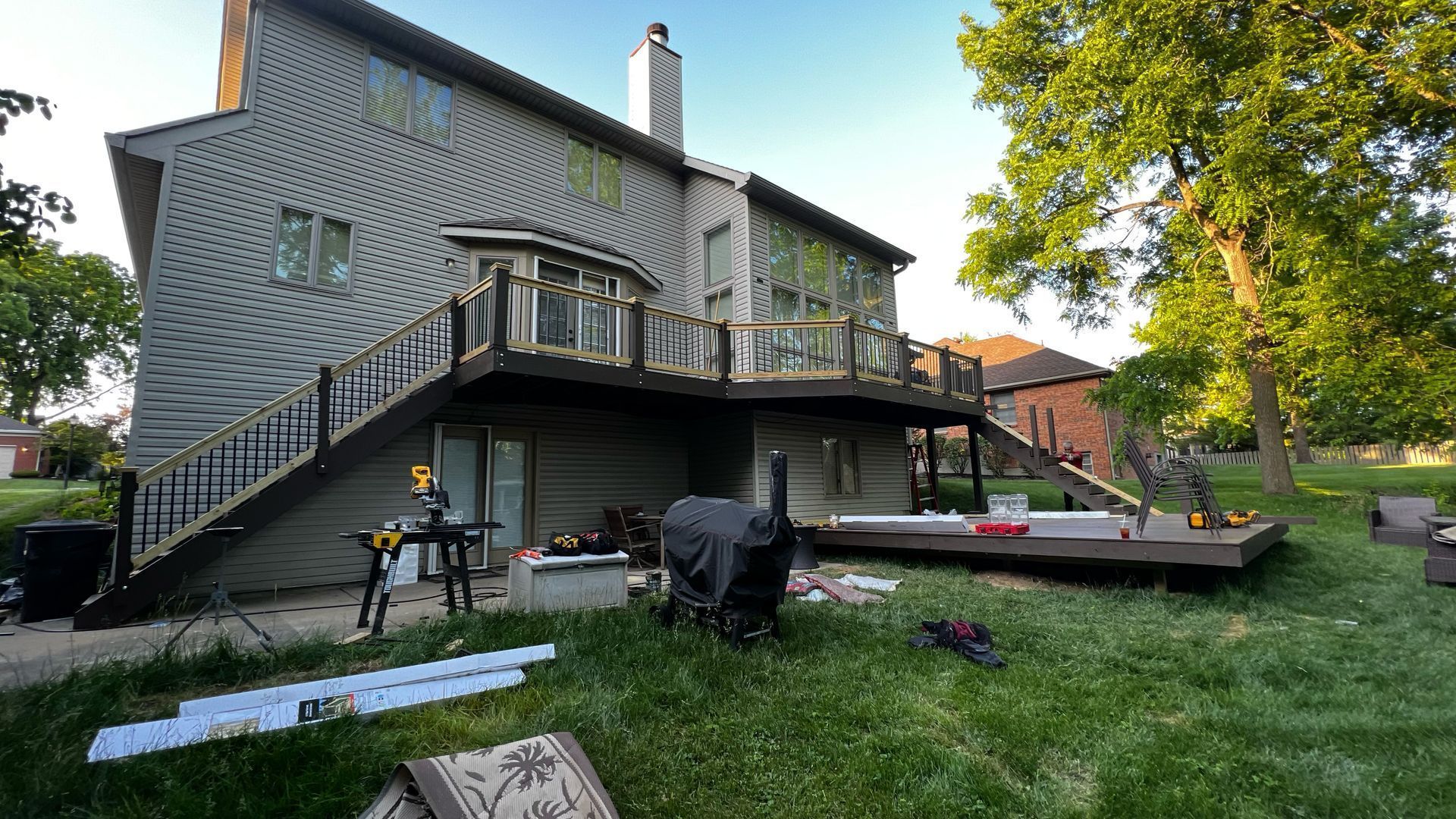 Backyard patio beside a gray house with deck, lawn chairs, grill, and trees in warm evening light