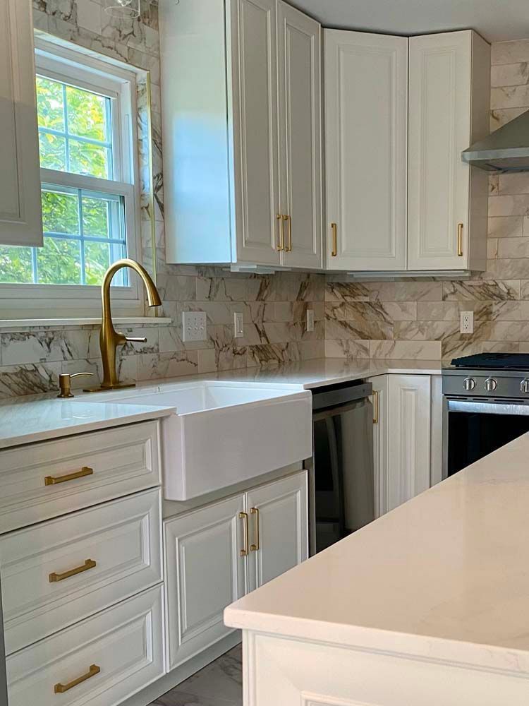 Bright white kitchen with marble countertops, gold faucet, and large window over the sink.