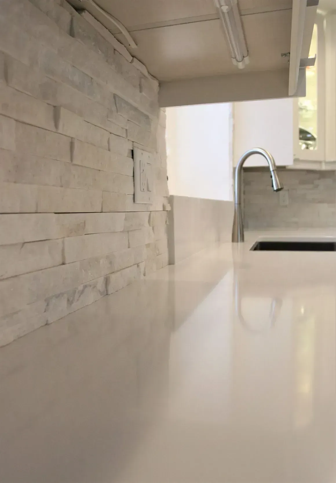 Modern kitchen with glossy white countertop, stone accent wall, and chrome sink faucet.