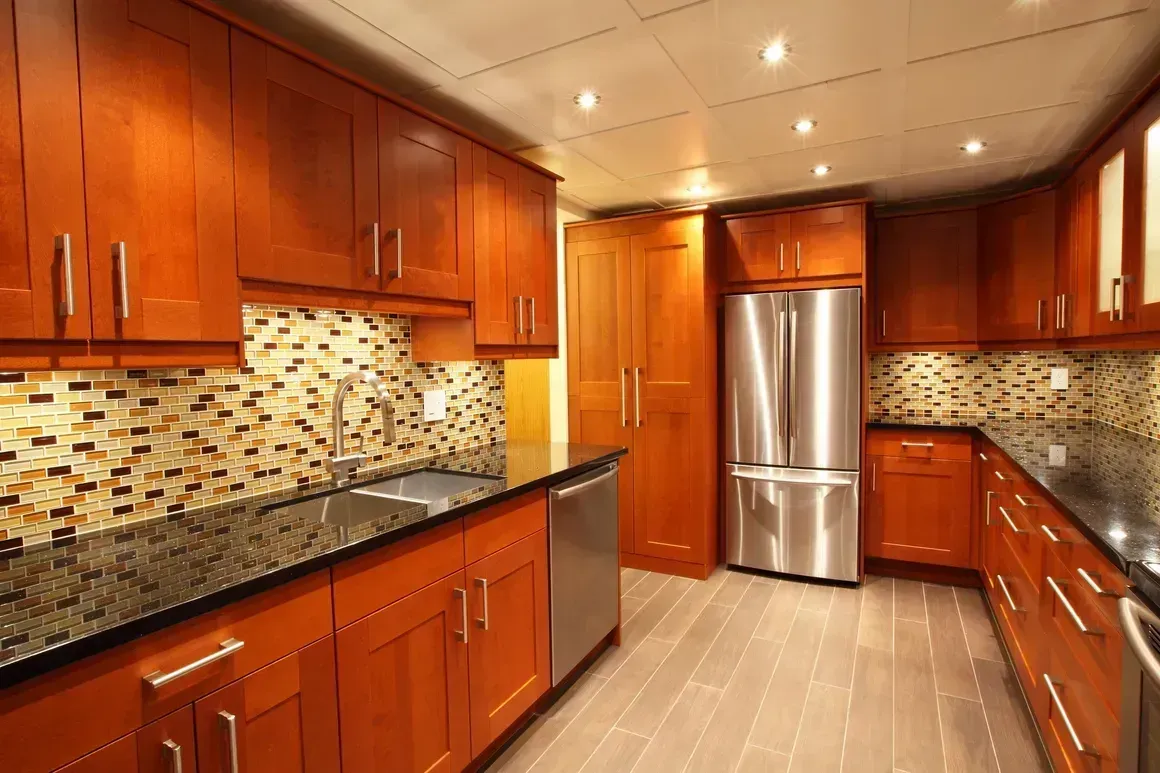 Modern kitchen with cherry cabinets, mosaic backsplash, black countertops, and stainless-steel refrigerator.
