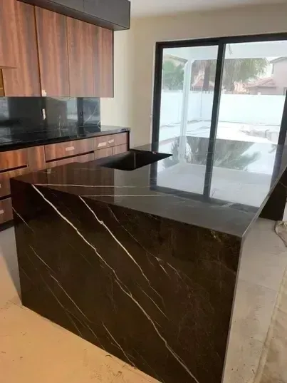 Modern kitchen with dark marble island, wood cabinets, and large sliding glass doors opening to a pool.