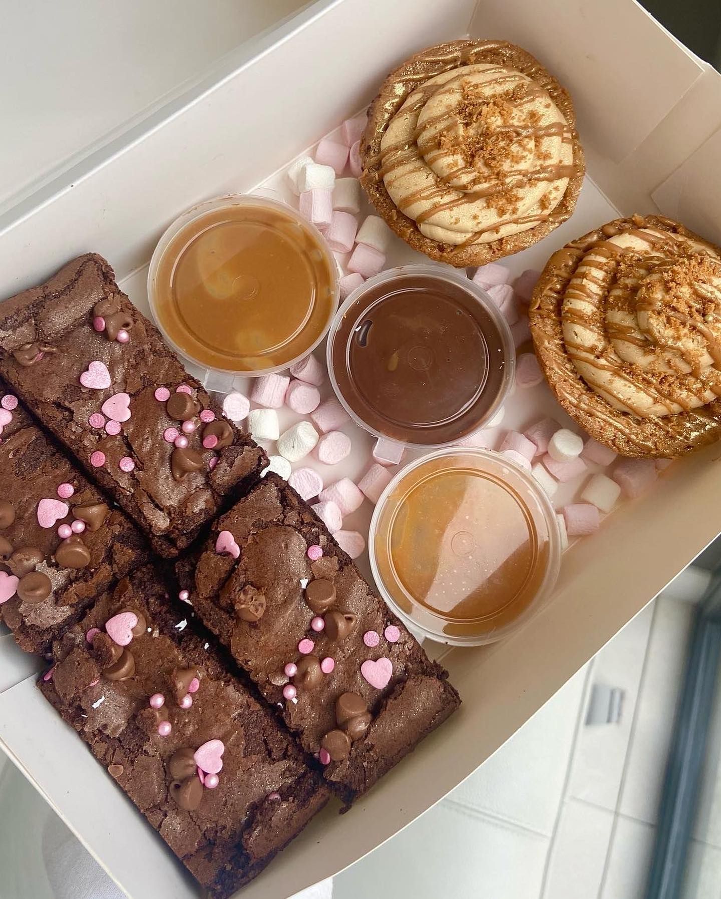 A box filled with brownies , cupcakes , sauces and marshmallows.