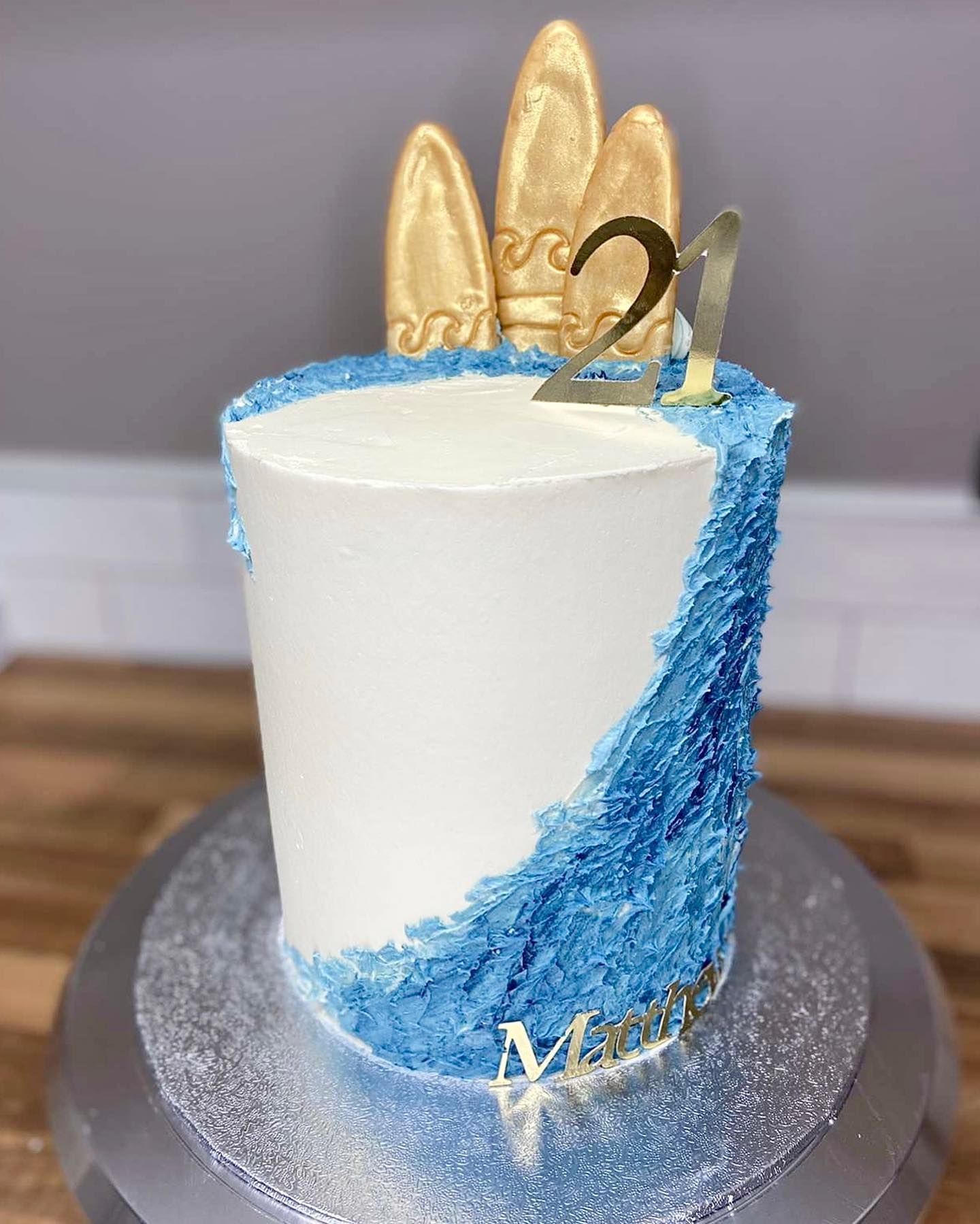A white cake with blue frosting and gold surfboards on top is on a cake stand.