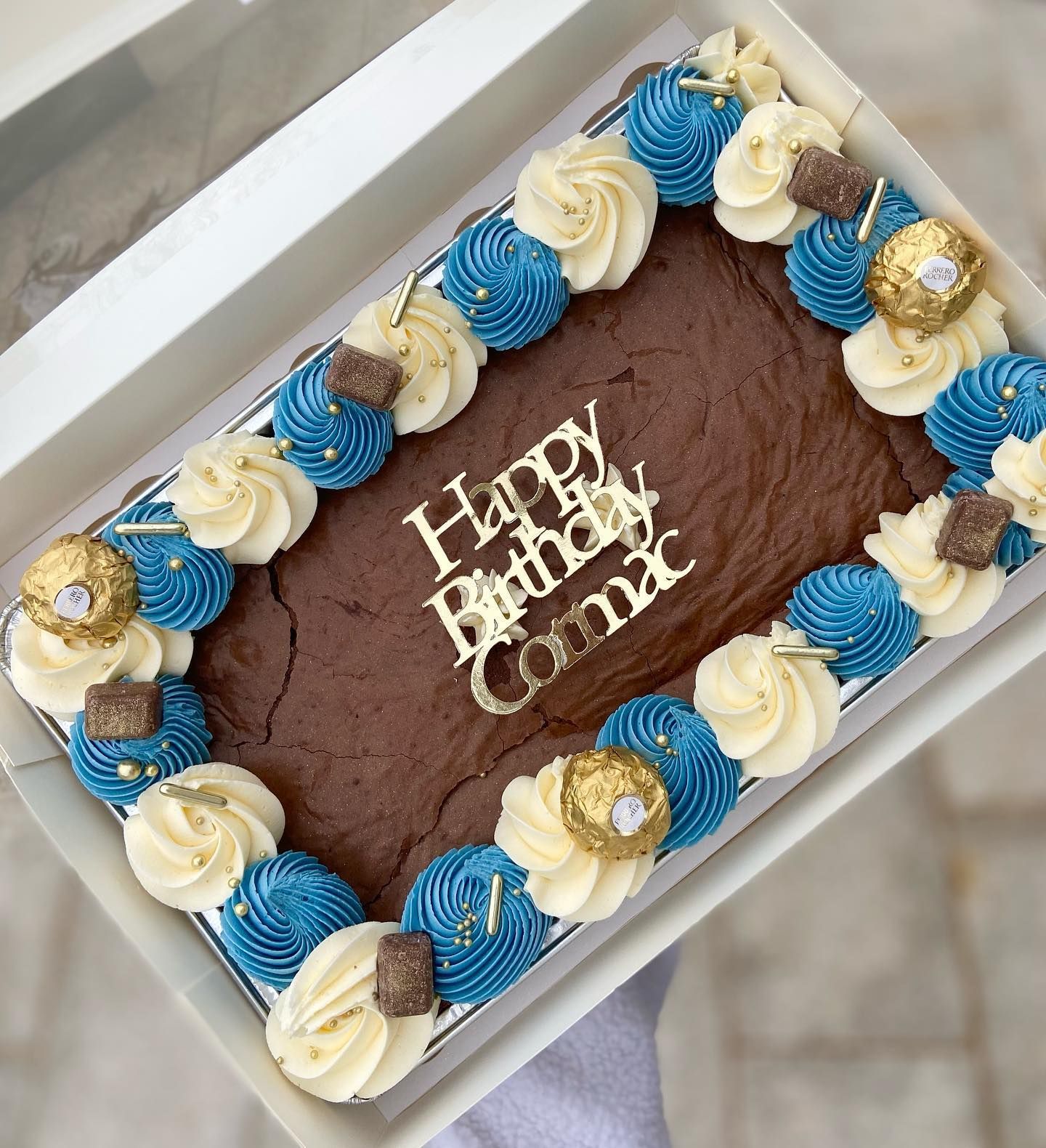 A brown cake with blue and white frosting that says happy birthday comac